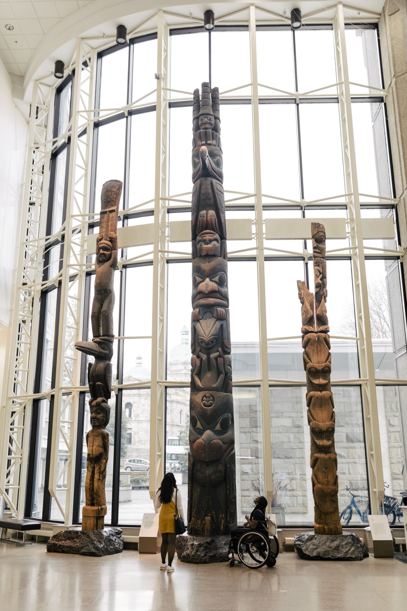 Two people at the Royal BC Museum, one standing, one in a wheelchair, gaze up at a collection of totem polls in front of a large bay window.