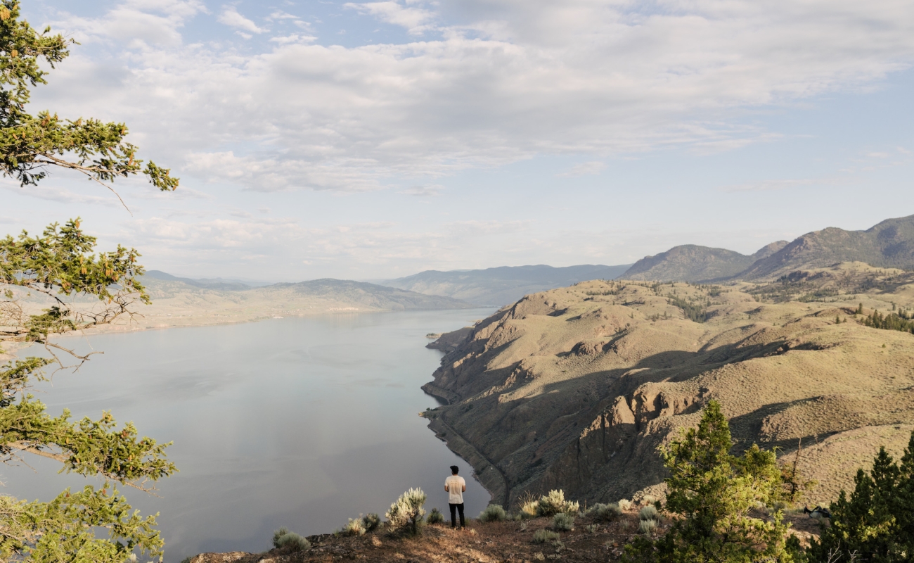 Una persona está de pie en un mirador rocoso, contemplando un lago vasto y tranquilo rodeado de colinas áridas y escarpadas. El paisaje se extiende a lo lejos, con montañas que se desvanecen en el horizonte bajo un cielo parcialmente nublado. Escasa vegetación y algunos árboles enmarcan el primer plano.