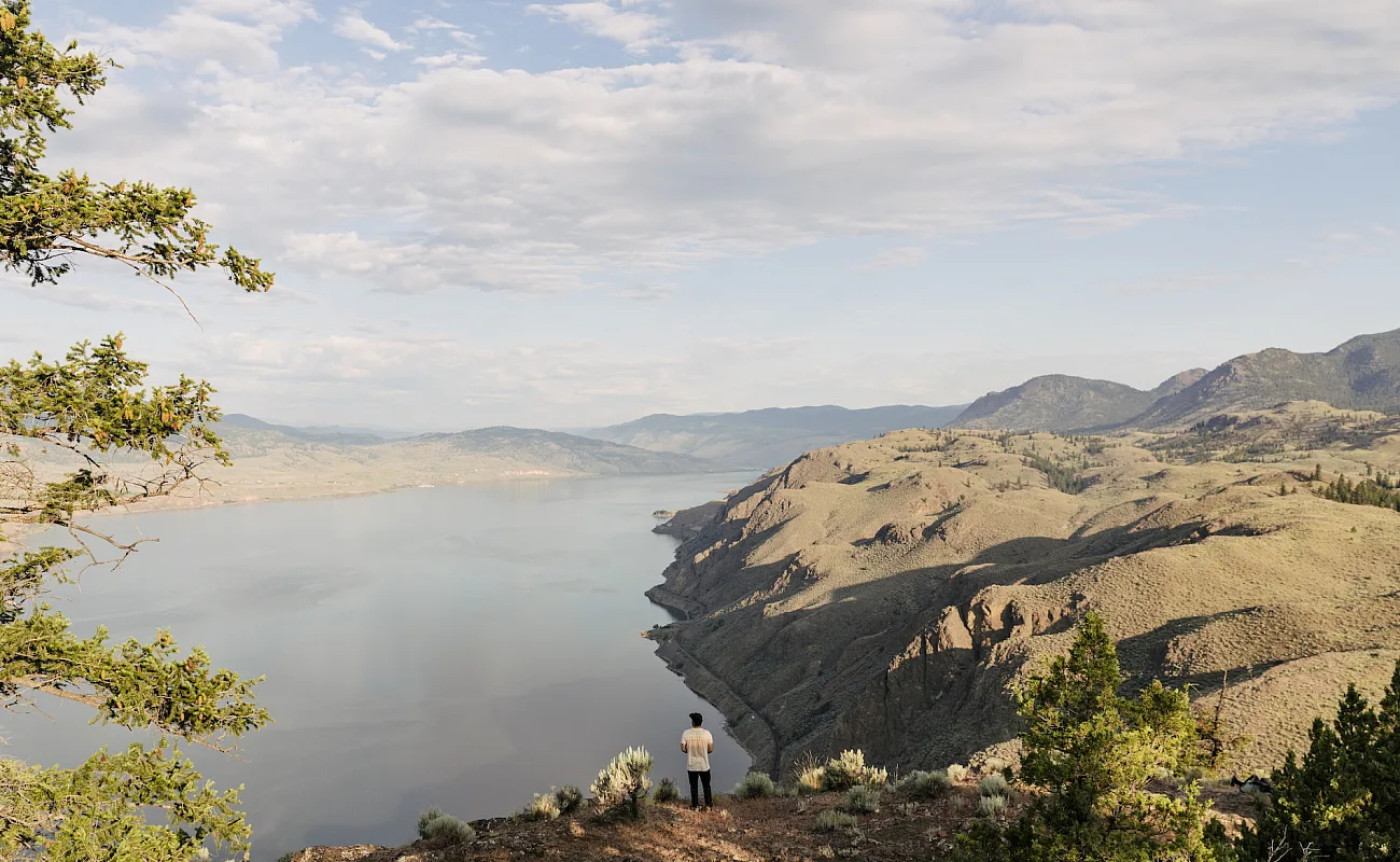 Eine Person steht auf einem felsigen Aussichtspunkt und blickt über einen weiten, ruhigen See, umgeben von trockenen, zerklüfteten Hügeln. Die Landschaft erstreckt sich in die Ferne, wobei Berge am Horizont unter einem teilweise bewölkten Himmel verschwimmen. Spärliche Vegetation und einige Bäume umrahmen den Vordergrund.