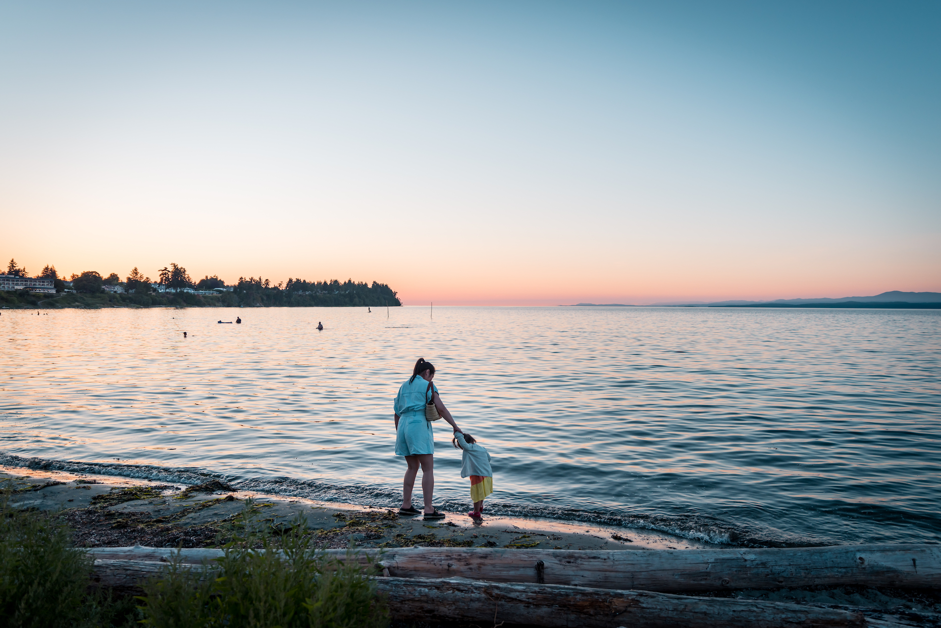 A mother and child walk along the beach in Parksville as the sunsets over the water. There are logs behind them on the sandy beach.