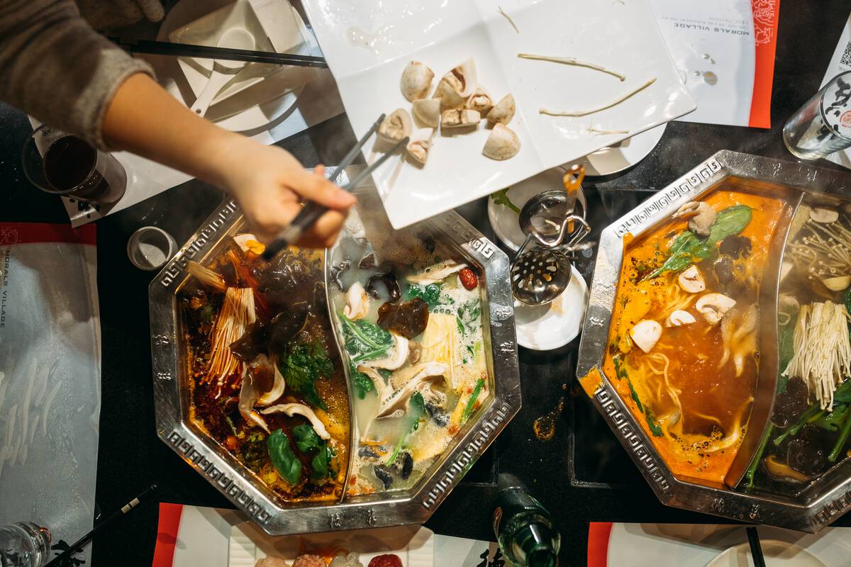 Food steams and simmers in who large, octagonal-shaped silver dishes, each divided in half. Above these dishes is a white platter with chopped mushrooms being placed into the steaming dishes with chopsticks.