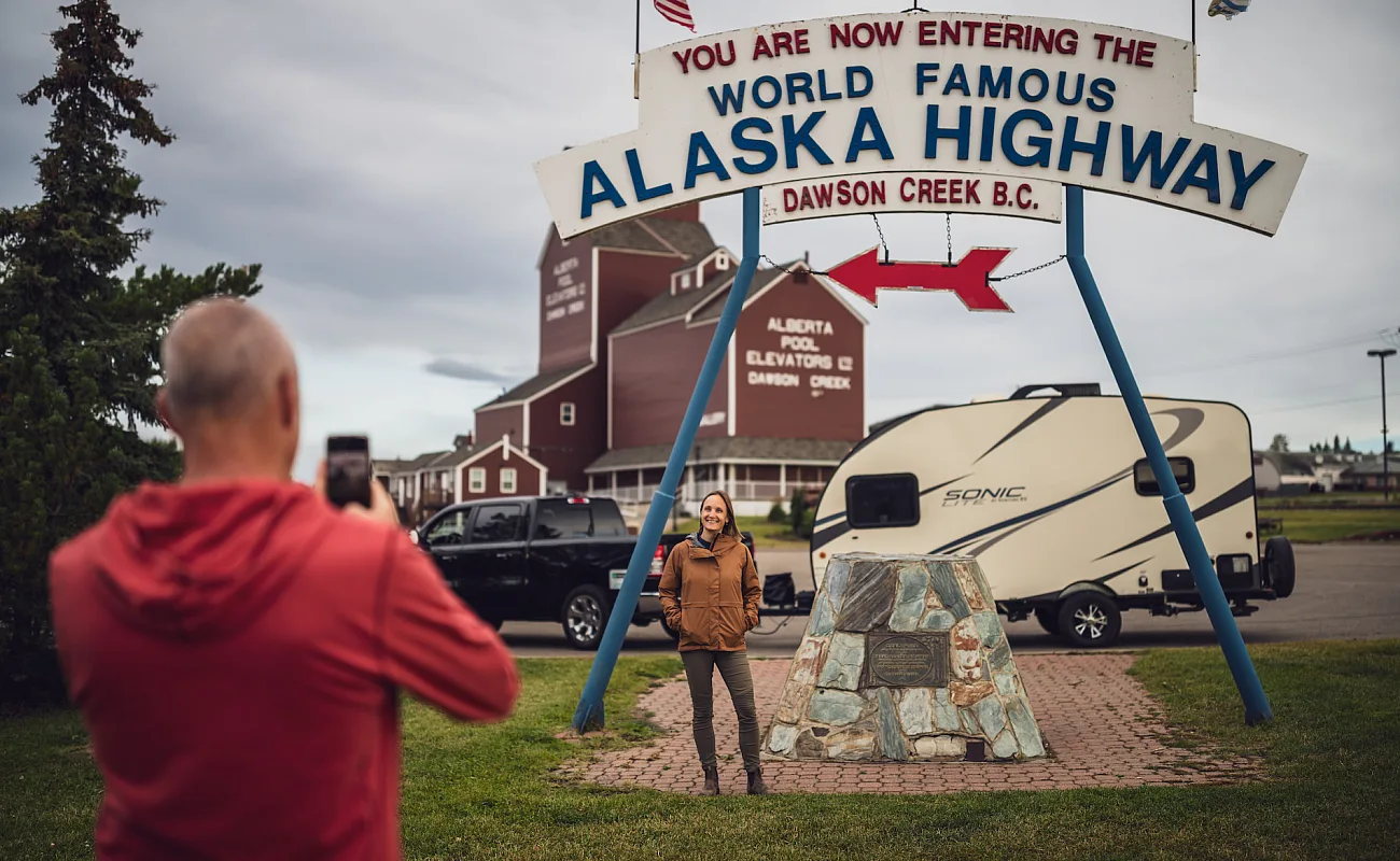 Un grupo de personas posando para una foto junto a un cartel que indica el punto kilométrico cero de la autopista Alaska Highway Dawson Creek, Columbia Británica.