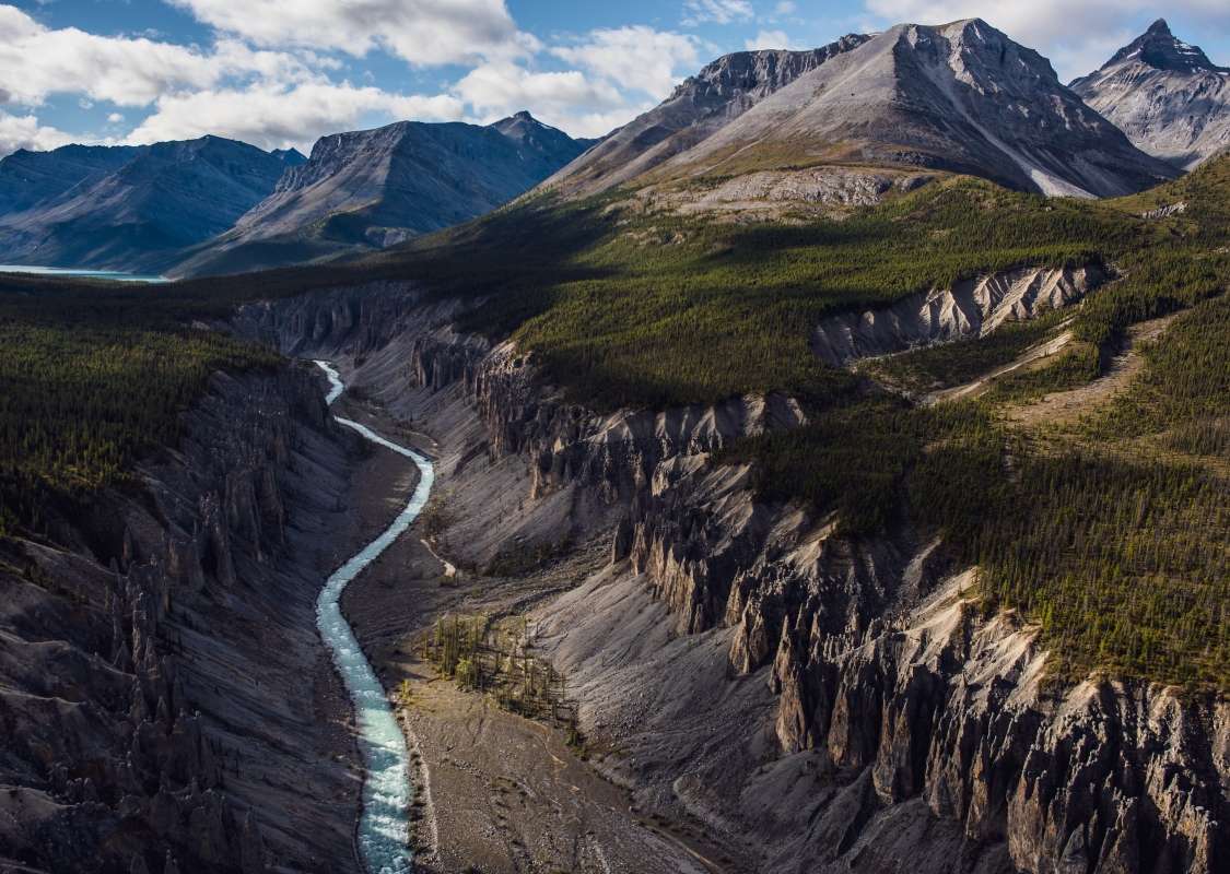 A torquoise river slices through a canyon edged by rocky cliffs, forested foothills and towering mountains in the background.