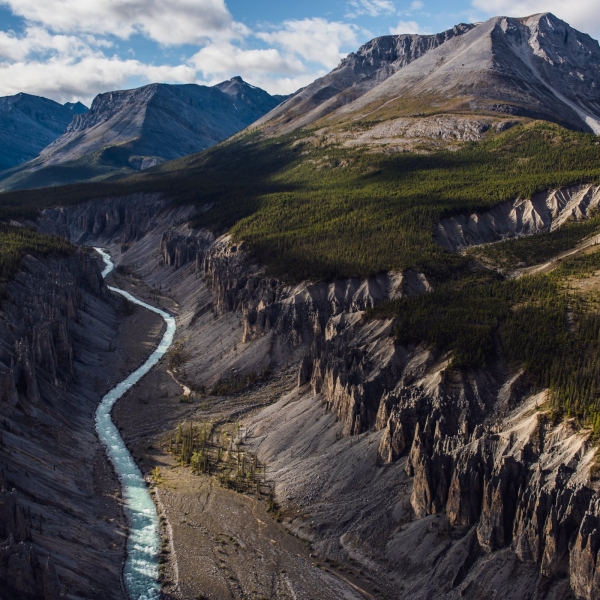 A torquoise river slices through a canyon edged by rocky cliffs, forested foothills and towering mountains in the background.