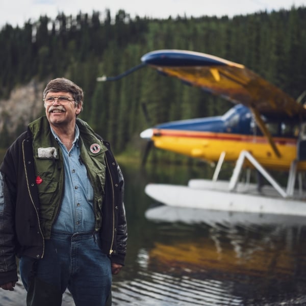 Ein robust aussehender Pilot geht neben seinem Wasserflugzeug, das auf einem abgelegenen See gelandet ist.