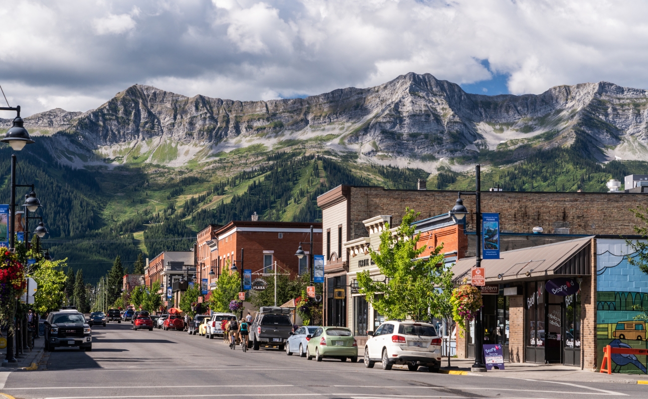 Cars are parked along a quiet street lined with heritage buildings. The sky is cloudy, and a dramatis mountain range is visible in the backdrop of the town.