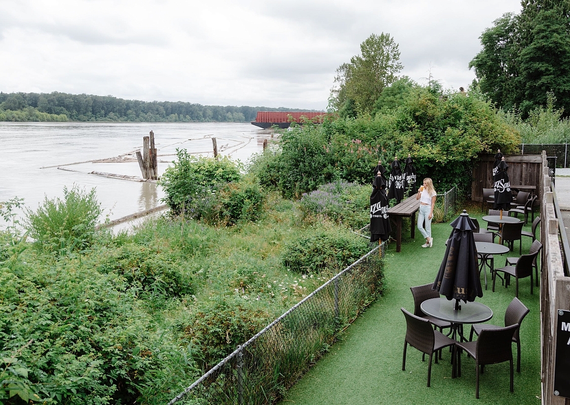 A person is standing at an outdoor patio on the waterfront filled with green shrubs and trees on an overcast day.