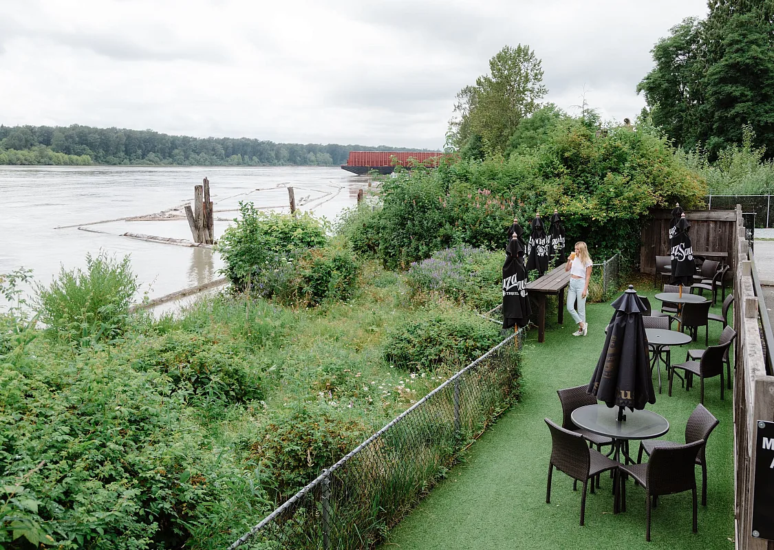 A person is standing at an outdoor patio on the waterfront filled with green shrubs and trees on an overcast day.
