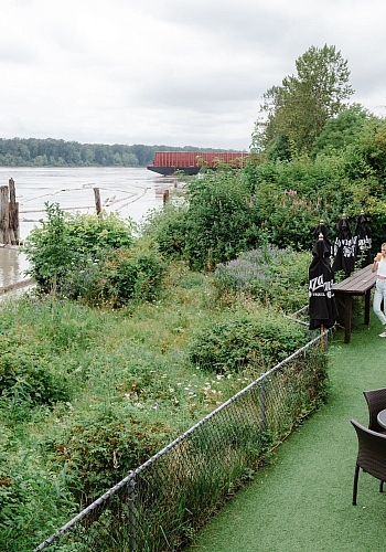 A person is standing at an outdoor patio on the waterfront filled with green shrubs and trees on an overcast day.