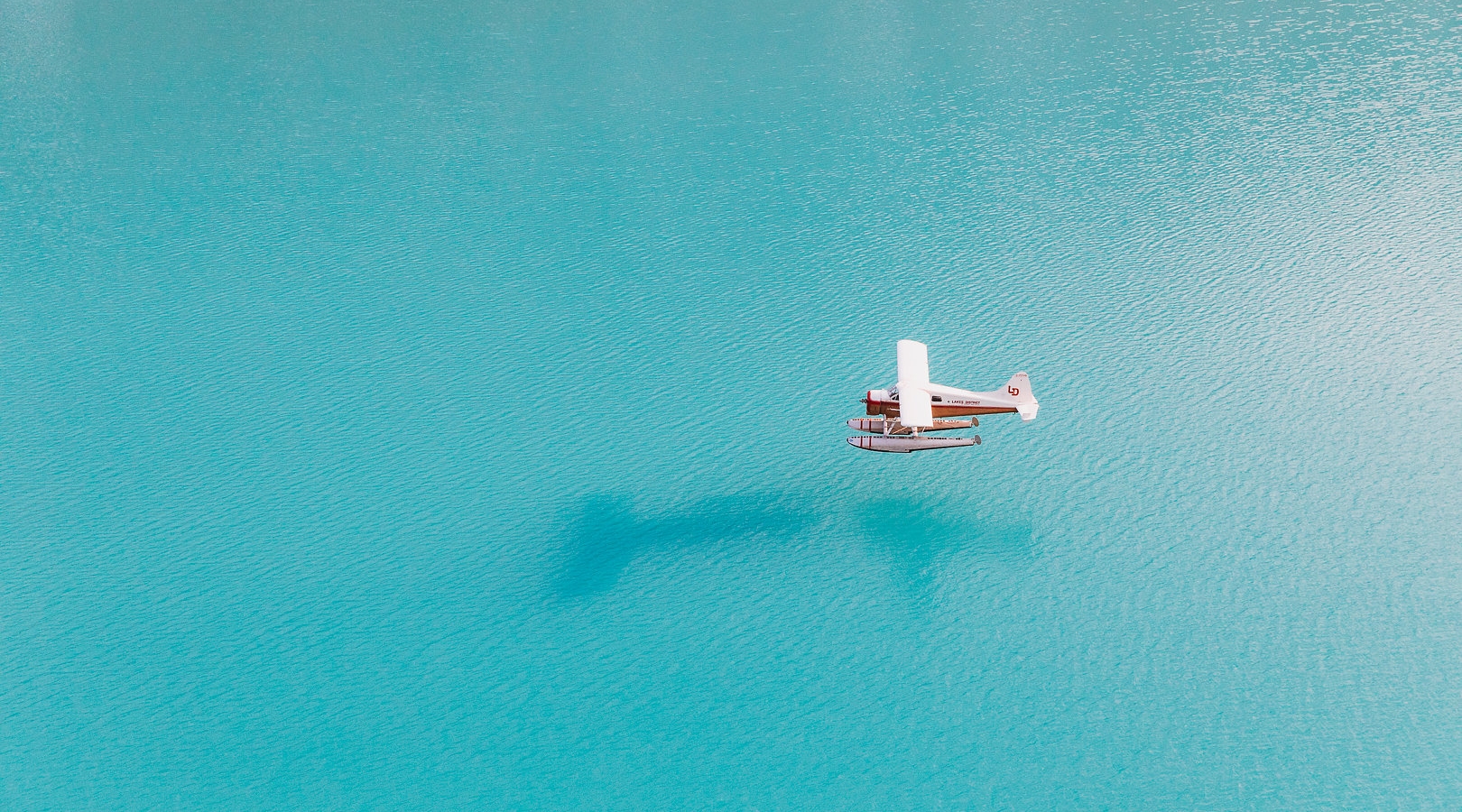 This still image is a placeholder for a video. A float plane above a glacier blue lake. The plane has white wings and a red body.