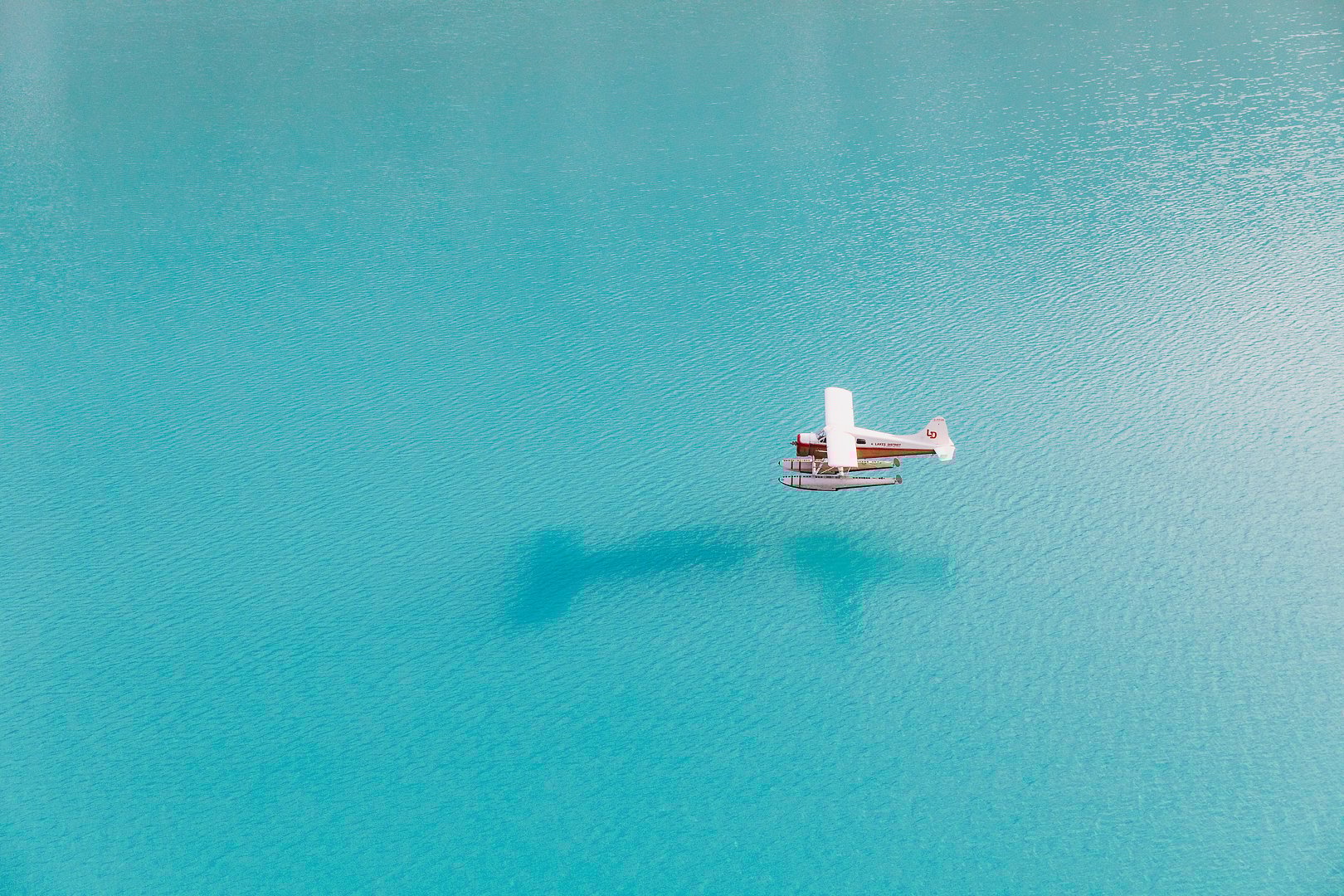 This still image is a placeholder for a video. A float plane above a glacier blue lake. The plane has white wings and a red body.