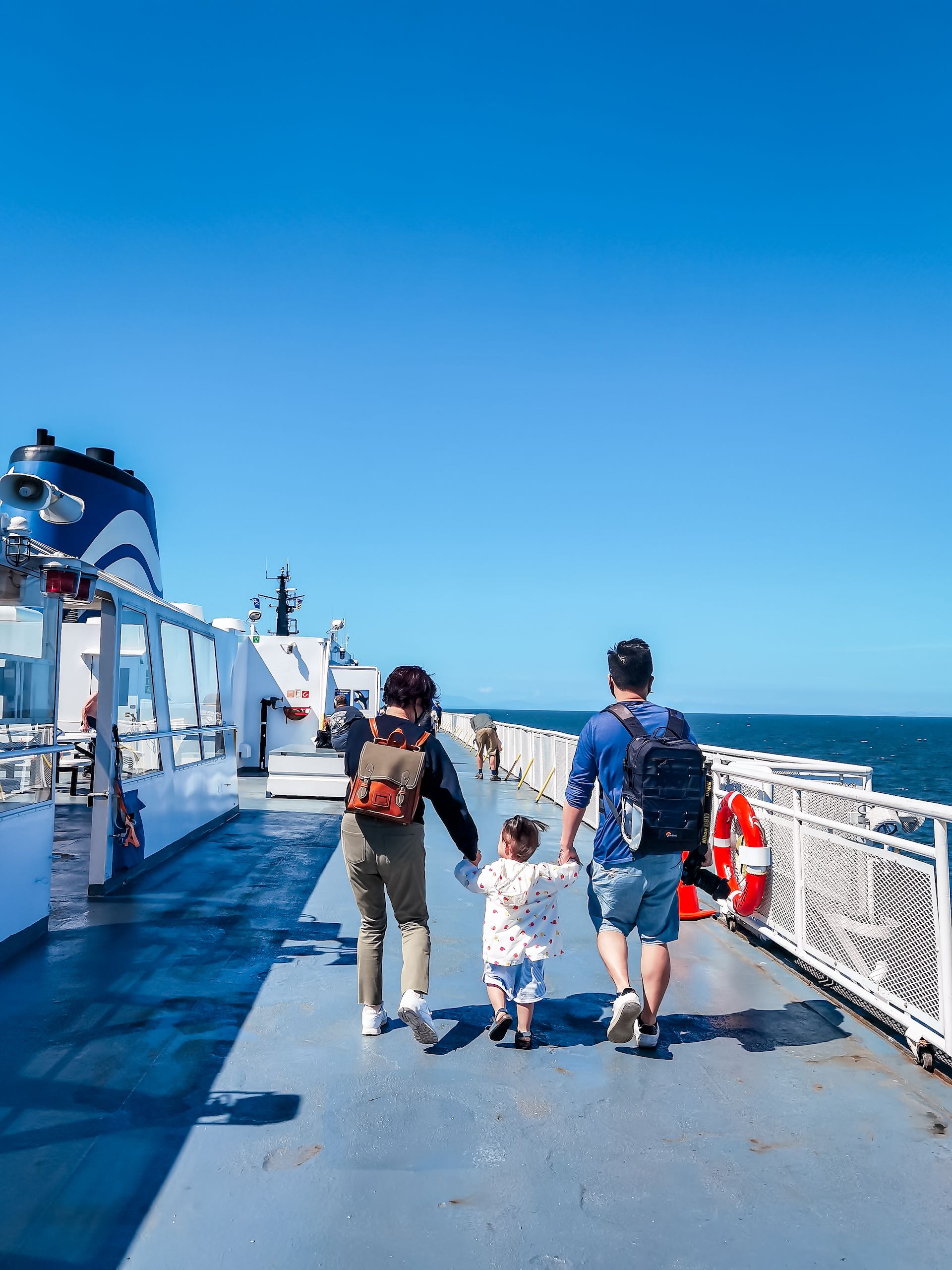 A family walks along the top deck of a BC Ferries boat on a sunny blue ski day. Two parents hold the hand of small child between them.