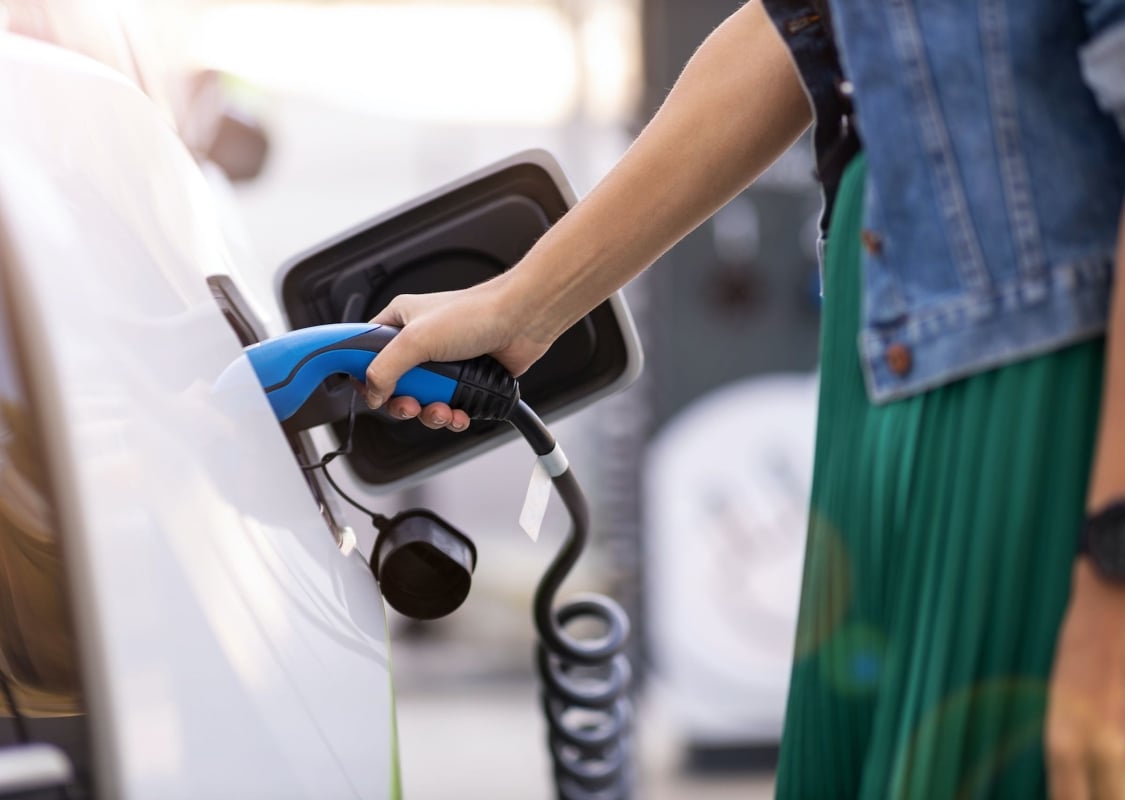 A white electric car is being charged by a women holding the handle. We see her arm and body only.