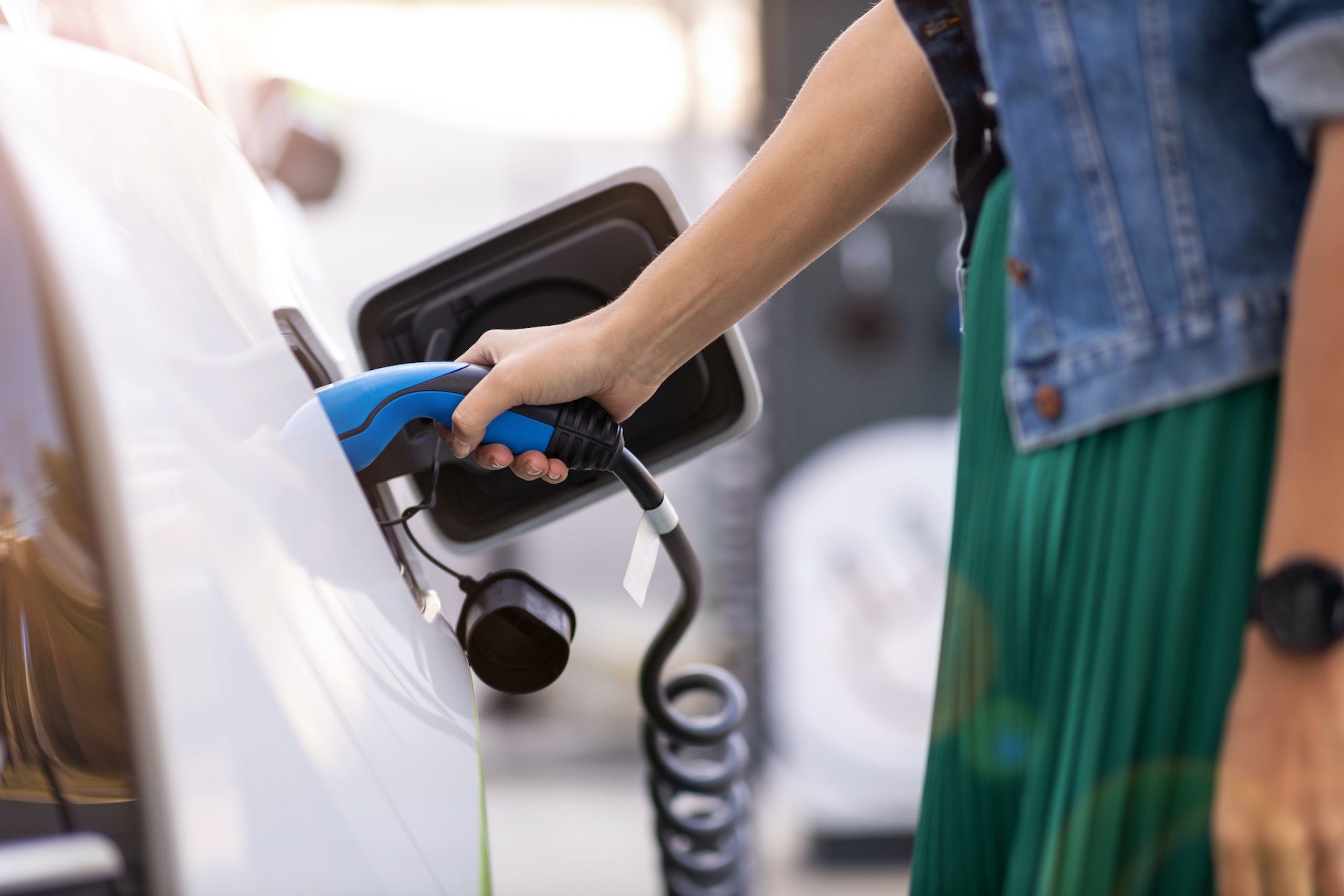 A white electric car is being charged by a women holding the handle. We see her arm and body only.
