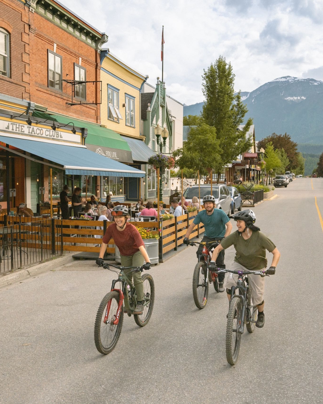 Tres amigos disfrutan de un paseo en bicicleta en el centro de Revelstoke mientras los locales comen algo en el patio de The Taco Club.