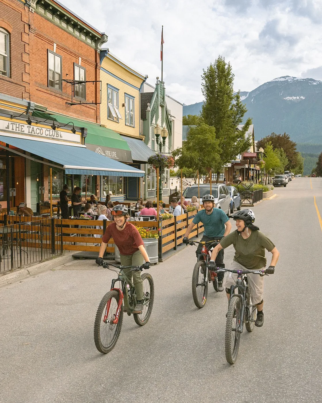 Tres amigos disfrutan de un paseo en bicicleta en el centro de Revelstoke mientras los locales comen algo en el patio de The Taco Club.