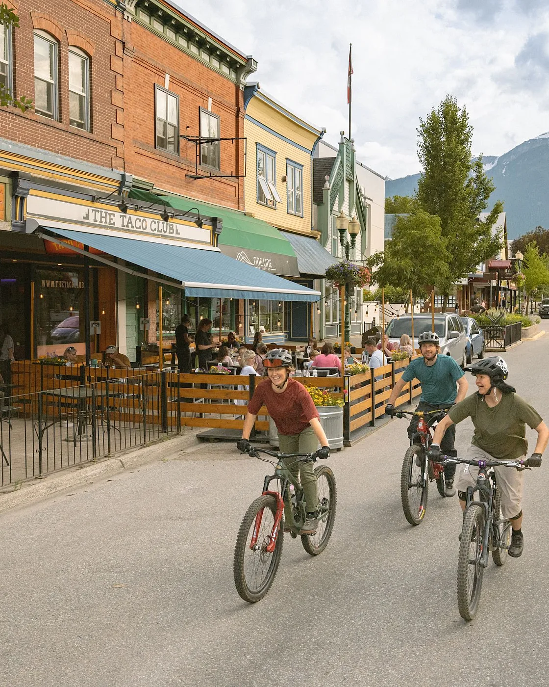 Tres amigos disfrutan de un paseo en bicicleta en el centro de Revelstoke mientras los locales comen algo en el patio de The Taco Club.