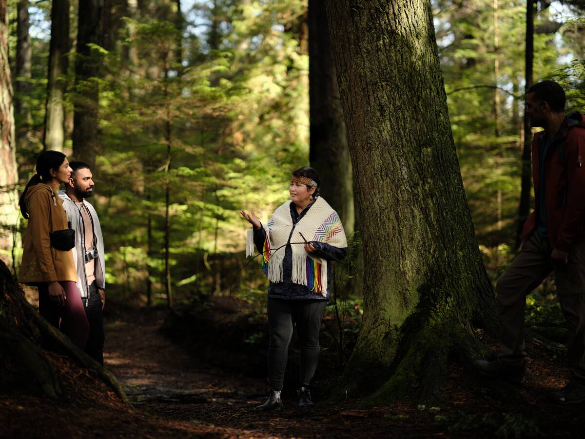 Candace Campo from Talaysay Walking Indigenous Tours is conducting a presentation and speaking to a group while on a walking experience in Stanley Park in Vancouver.