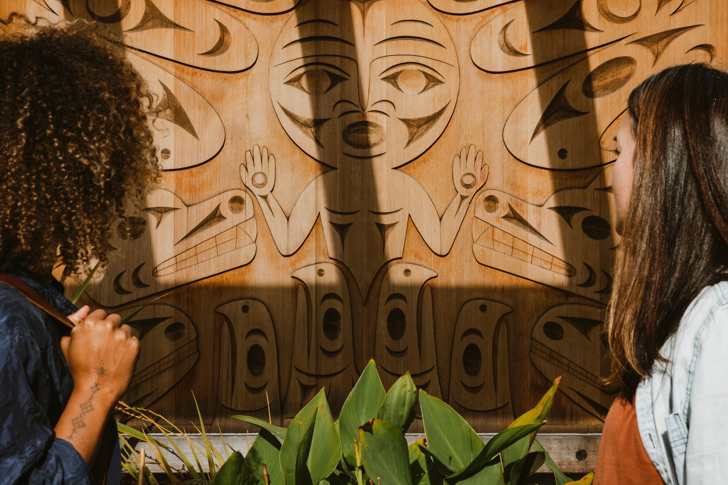 Two people taking in an Indigenous carving on the exterior wall at the Gibsons Public Art Gallery.
