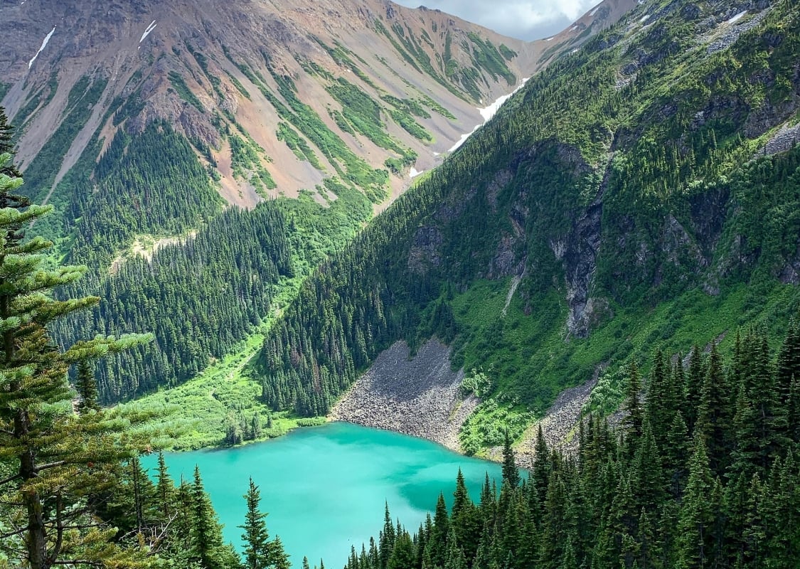 A colourful mountain scene centring around a vibrant blue lake that is surrounded by orange mineral mountains and flanked by by lush green forest.