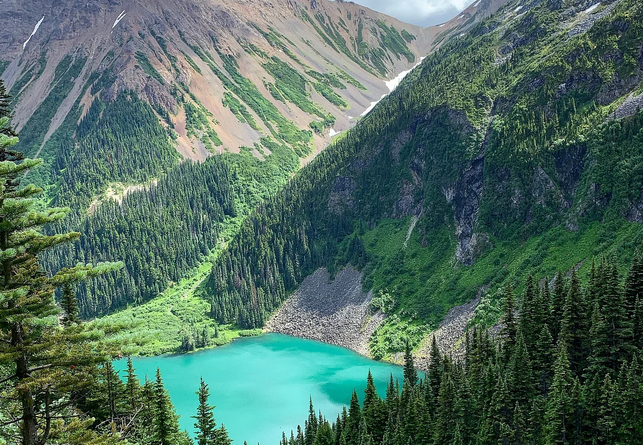 A colourful mountain scene centring around a vibrant blue lake that is surrounded by orange mineral mountains and flanked by by lush green forest.