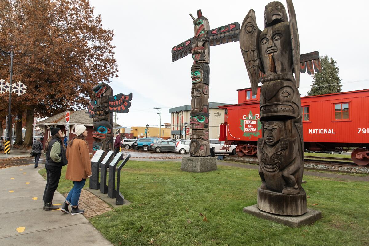Two people read signage and admire totem poles with a rail car in the background.