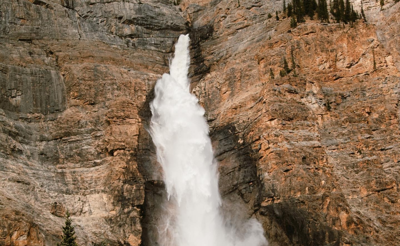 A waterfall plunges down a steep, rocky cliff face, surrounded by rugged terrain and pine trees.