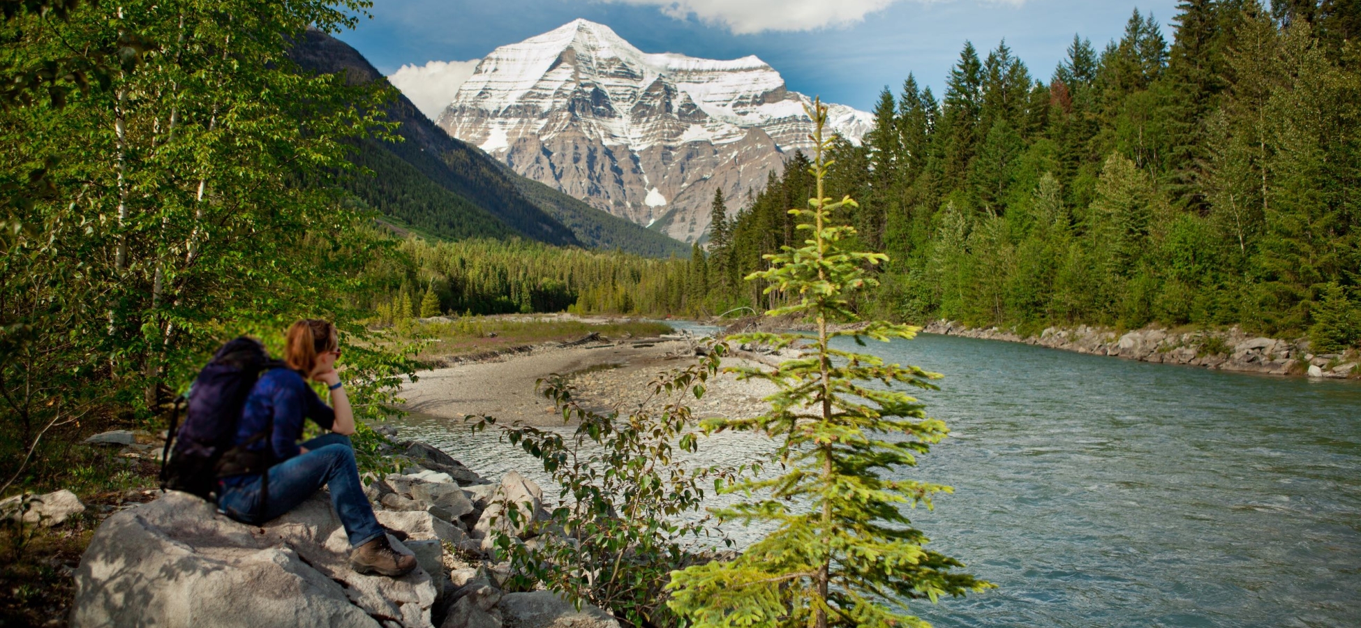 Eine Person sitzt auf einem Felsen neben einem Fluss und blickt auf Mount Robson.