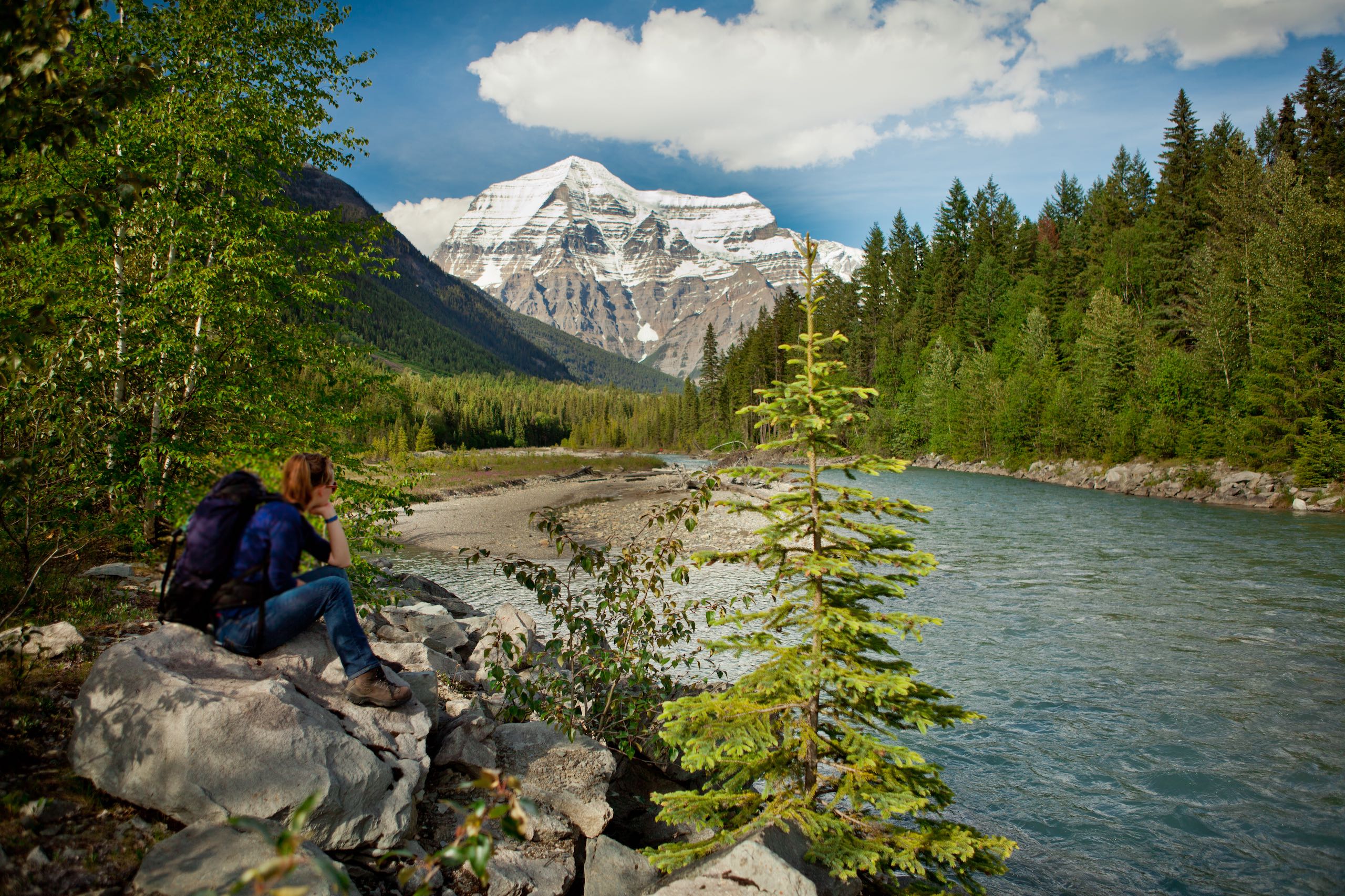 A person sitting on a rock next to a river looking at Mount Robson.