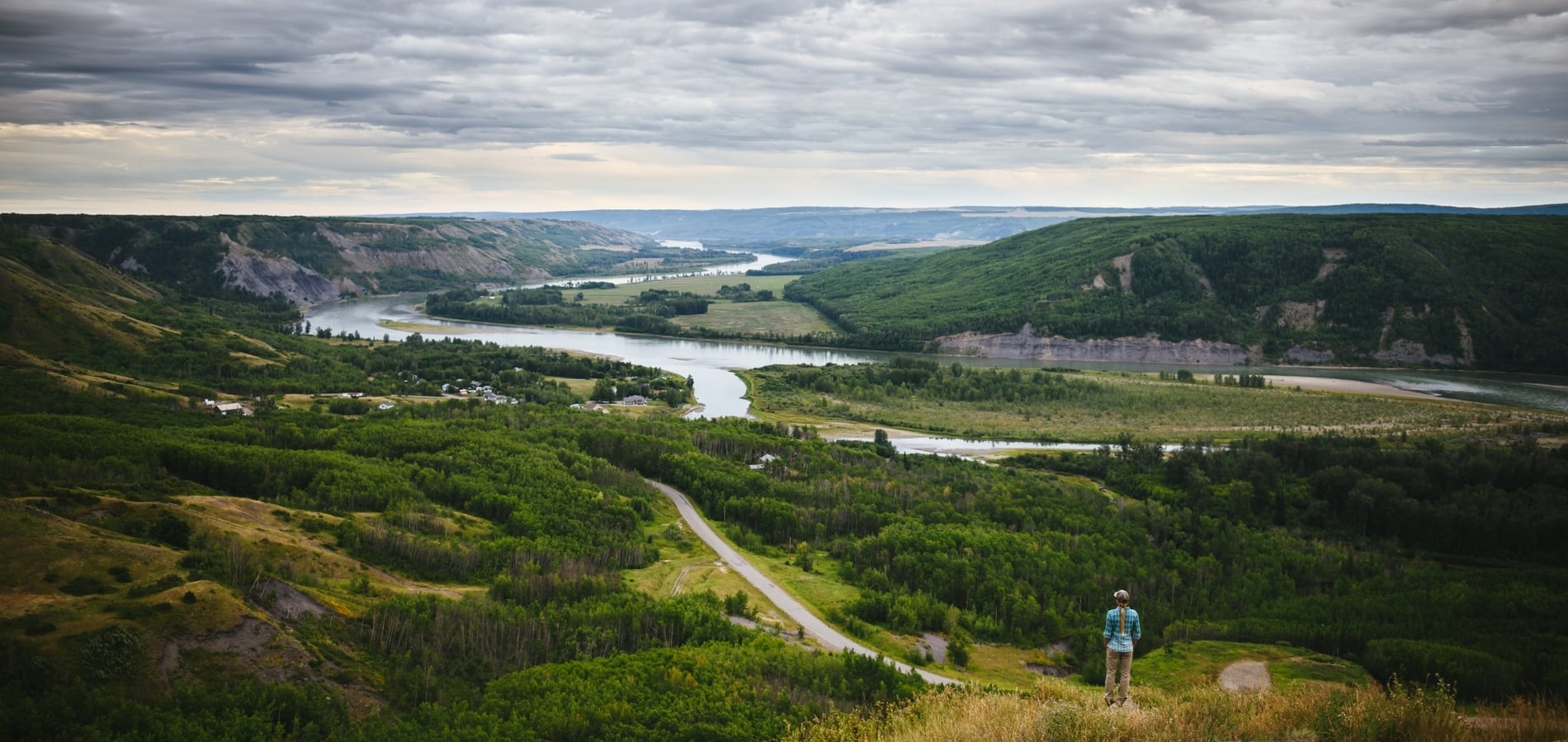A person in a blue jacket stand on a hill overlooking a fertile river valley
