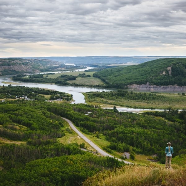 A person in a blue jacket stand on a hill overlooking a fertile river valley