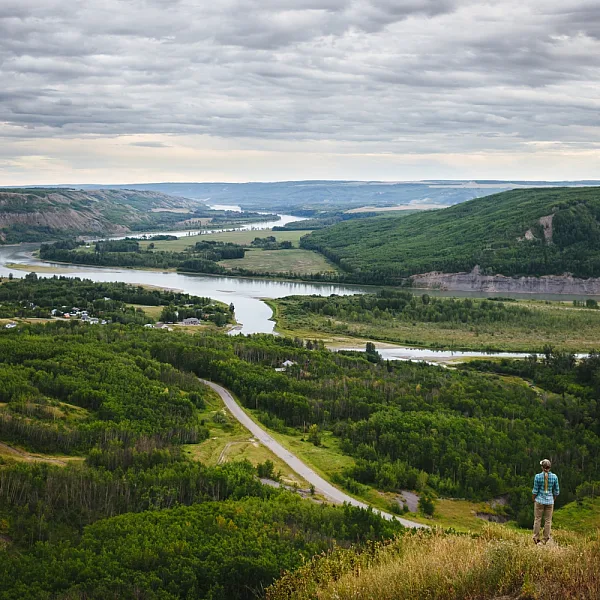 A person in a blue jacket stand on a hill overlooking a fertile river valley