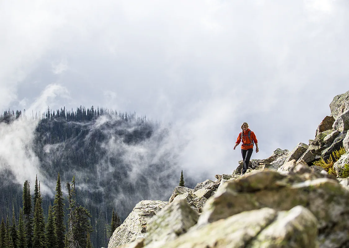 A hiker moves along a rocky ledge overlooking thick forest and fog.