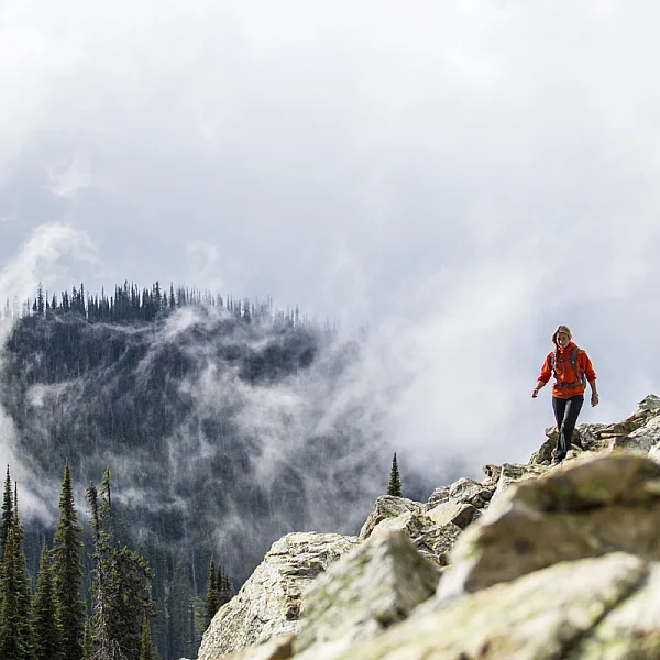 A hiker moves along a rocky ledge overlooking thick forest and fog.