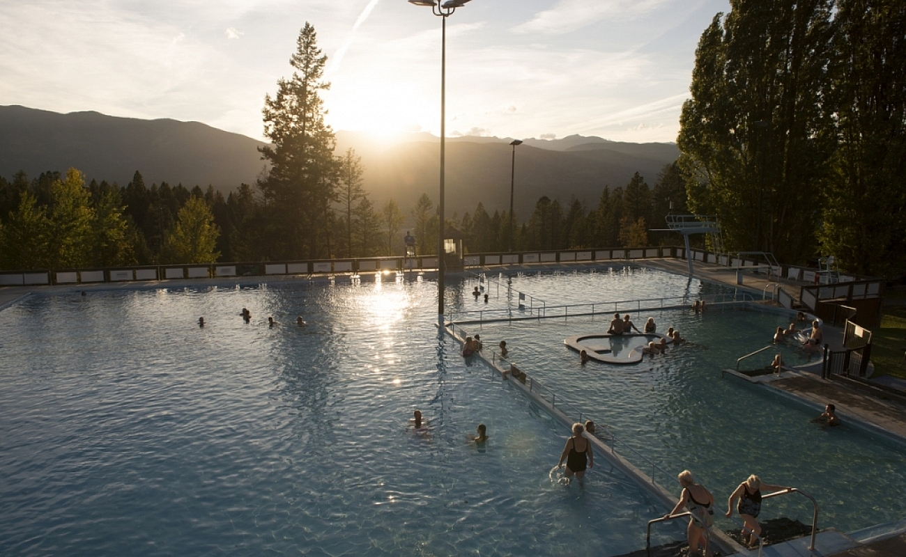 An aerial shot of a large hot springs pool with 20 or so people in the water. In the distance the sun is setting over the mountains and there are some evergreen trees just beyond the pool.