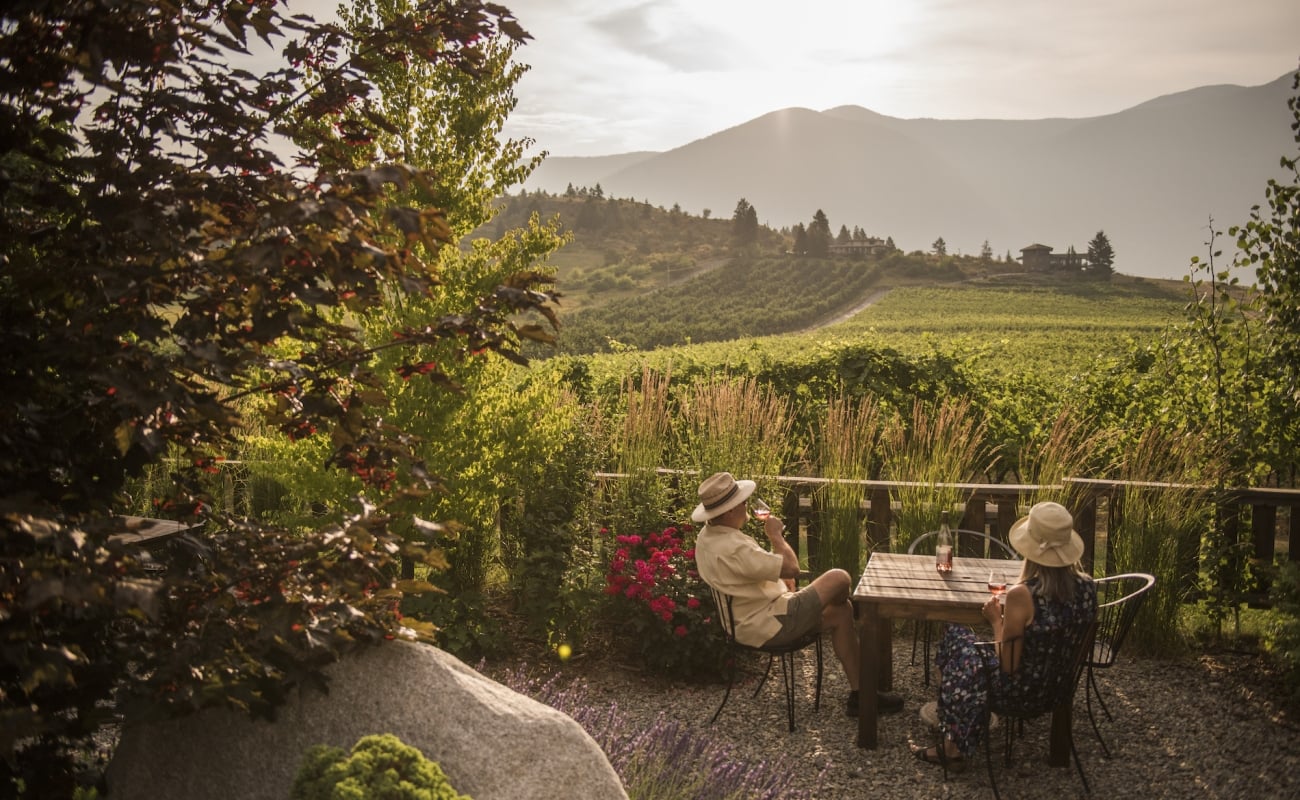 Two people sitting on a patio looking over a winery at a table.