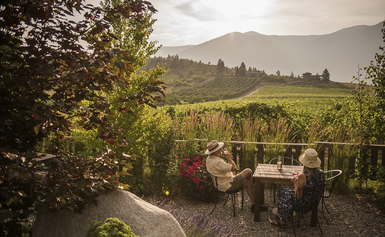Zwei Menschen sitzen auf einer Terrasse mit Blick auf ein Weingut an einem Tisch.