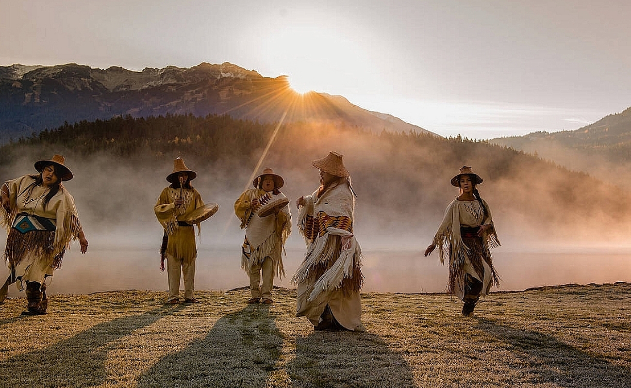 Five Indigenous people in traditional attire dance and play drums in a field with mist rising and the sun peeking over the mountains in the distance