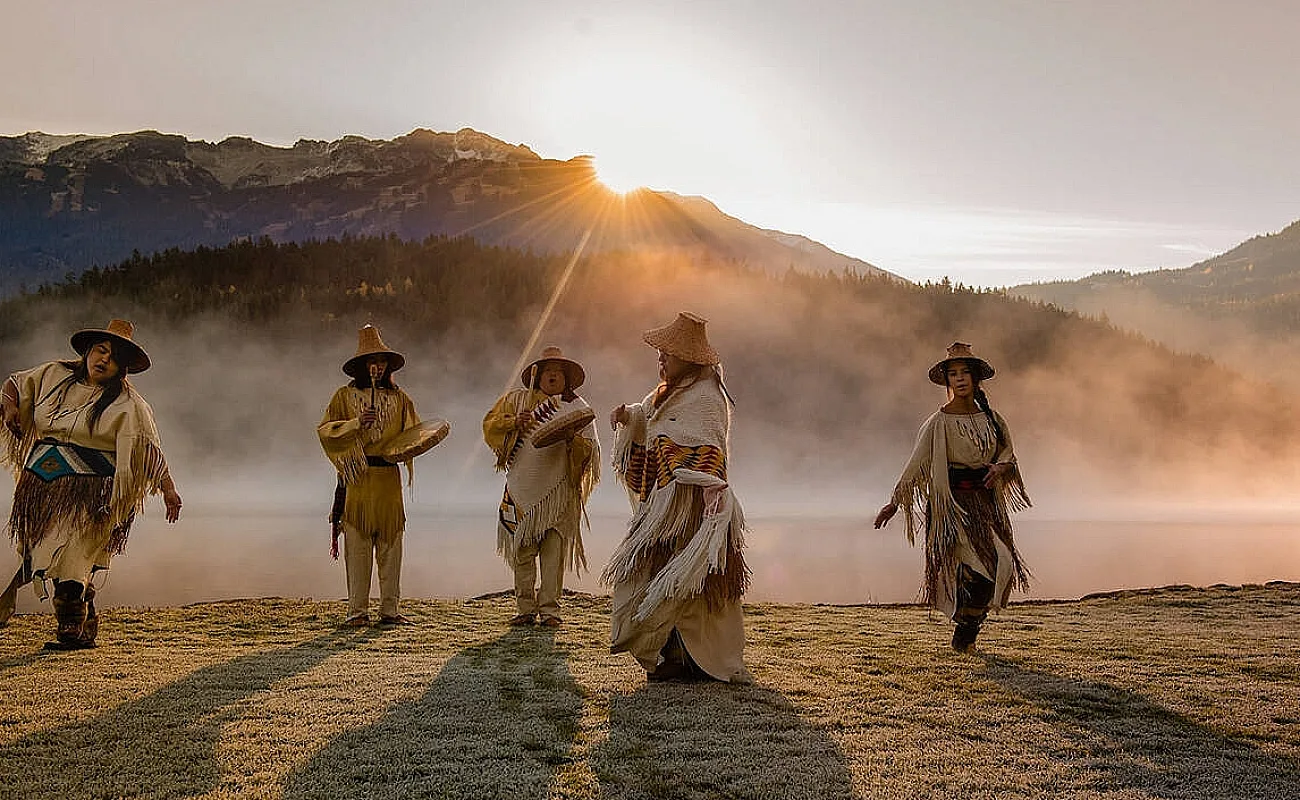 Five Indigenous people in traditional attire dance and play drums in a field with mist rising and the sun peeking over the mountains in the distance