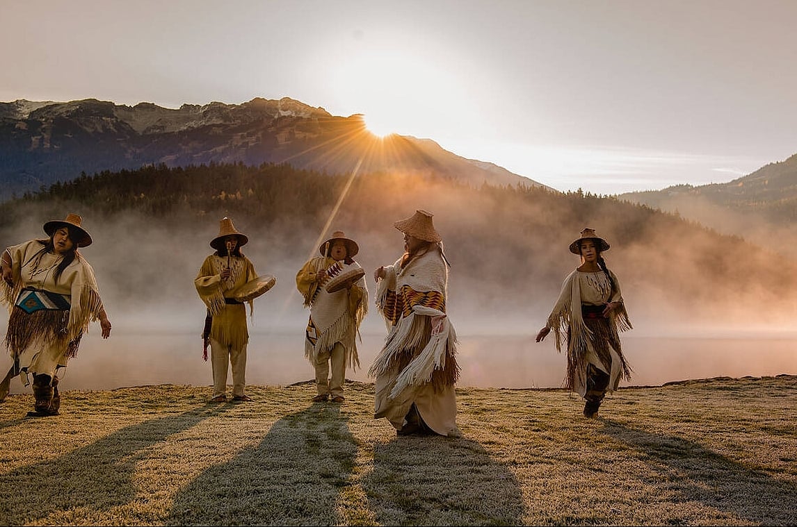 Five Indigenous people in traditional attire dance and play drums in a field with mist rising and the sun peeking over the mountains in the distance