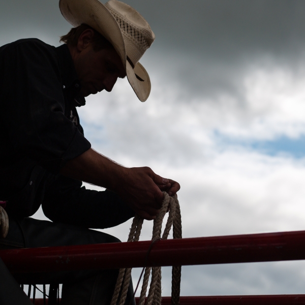 Silhouette of a cowboy in a hat sitting on a fence, holding a rope. Cloudy sky is in the background.