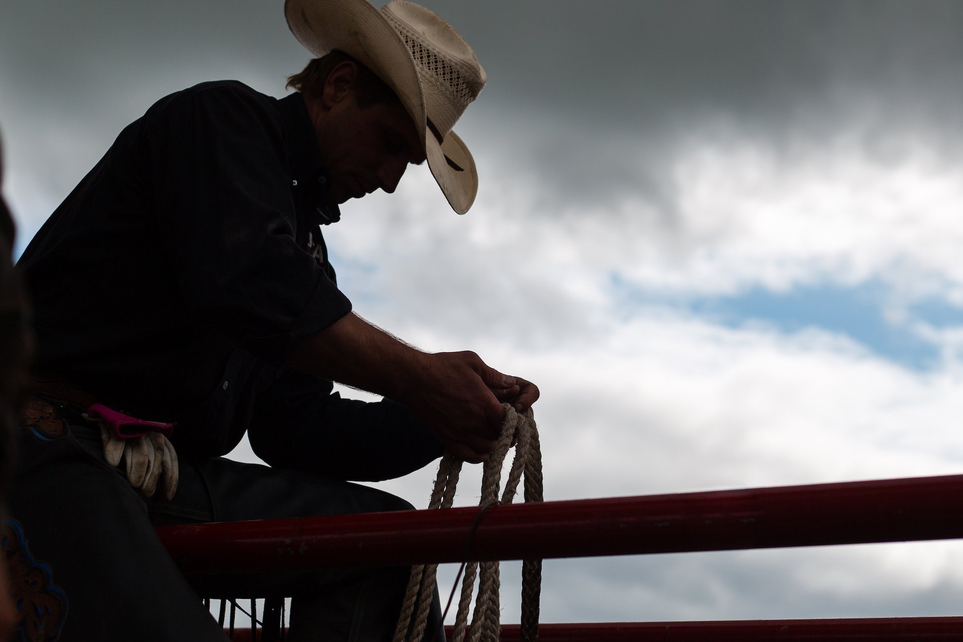 Silhouette of a cowboy in a hat sitting on a fence, holding a rope. Cloudy sky is in the background.