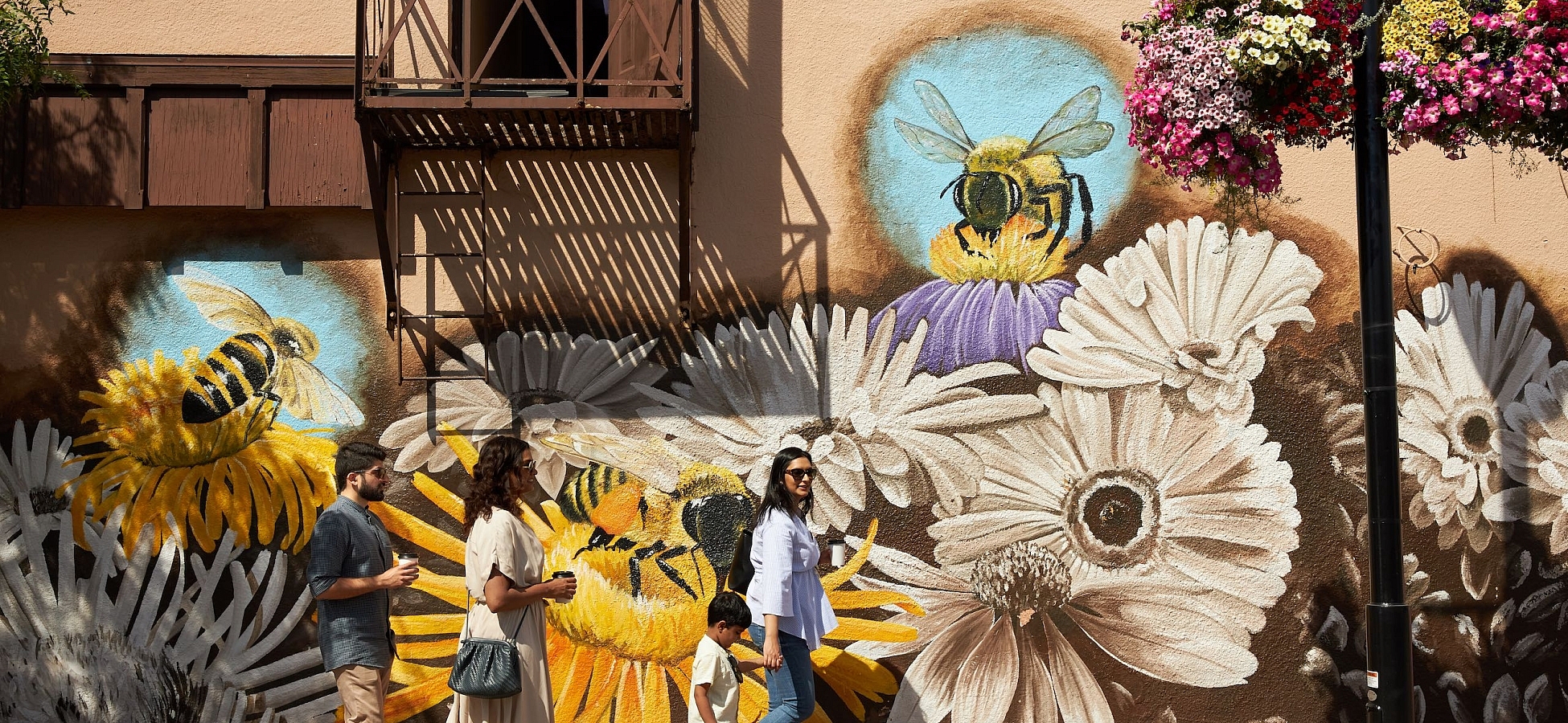Three people walk on a paved road with a colourful mural of white and yellow daisies and bees pollinating the flowers on the walls of the building behind them.