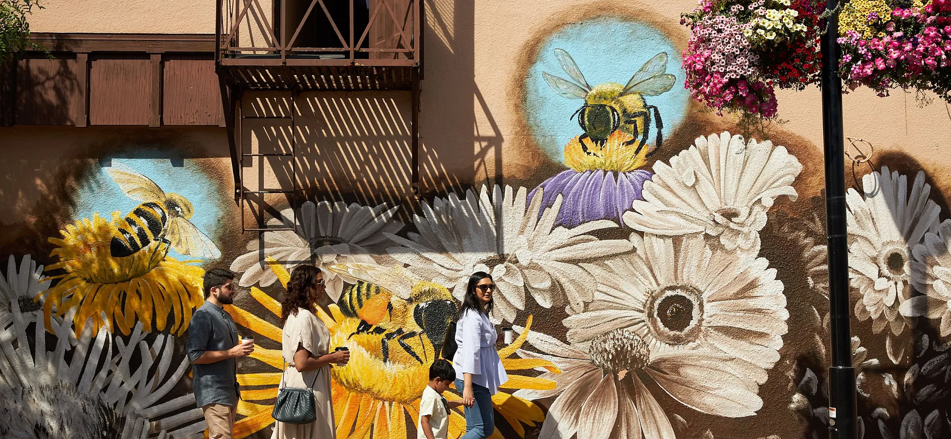 Three people walk on a paved road with a colourful mural of white and yellow daisies and bees pollinating the flowers on the walls of the building behind them.