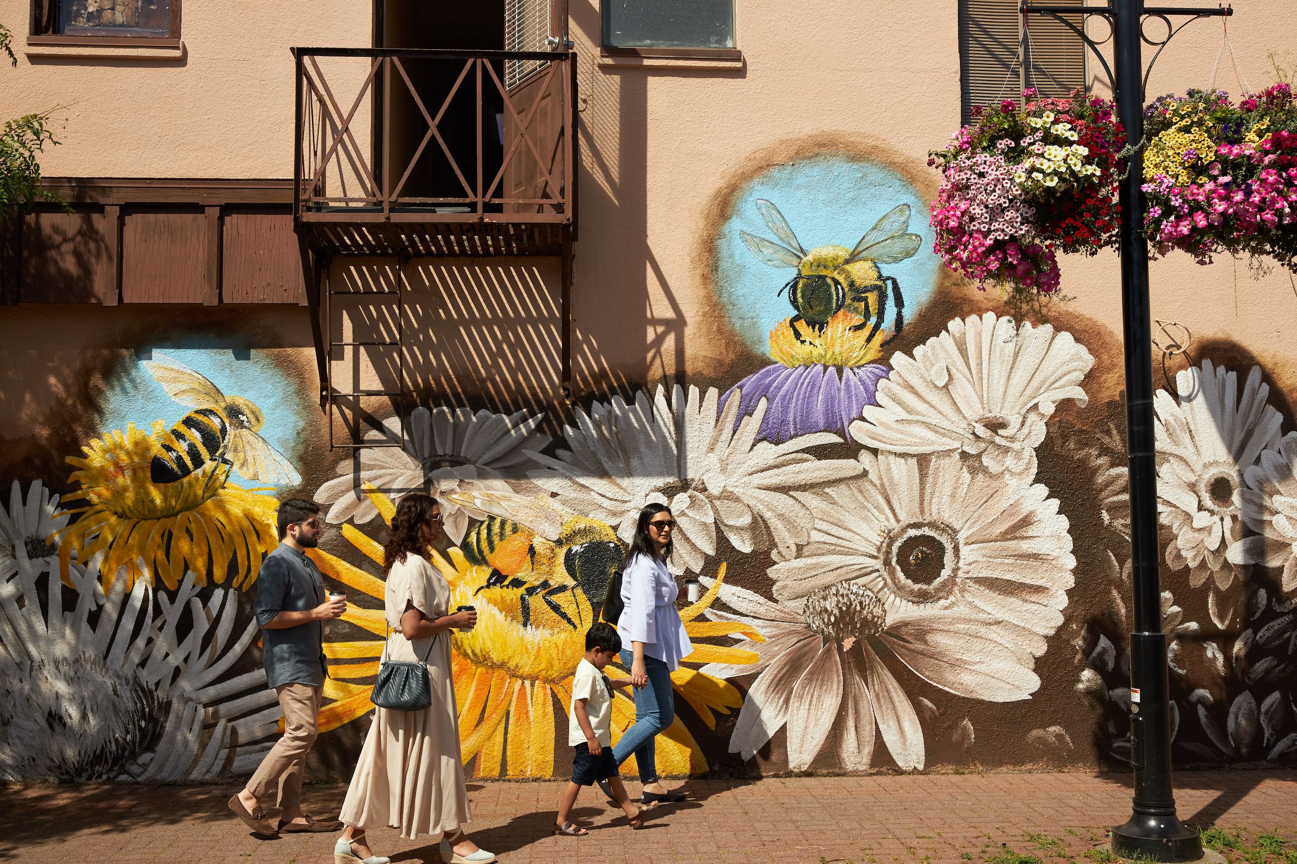 Three people walk on a paved road with a colourful mural of white and yellow daisies and bees pollinating the flowers on the walls of the building behind them.