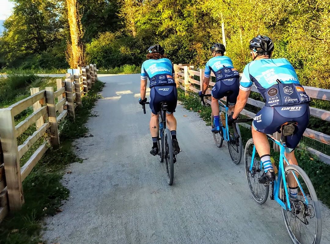 Three cyclists on turquoise bikes riding along a curved gravel path lined by wooden fences on each side with trees ahead of the path.