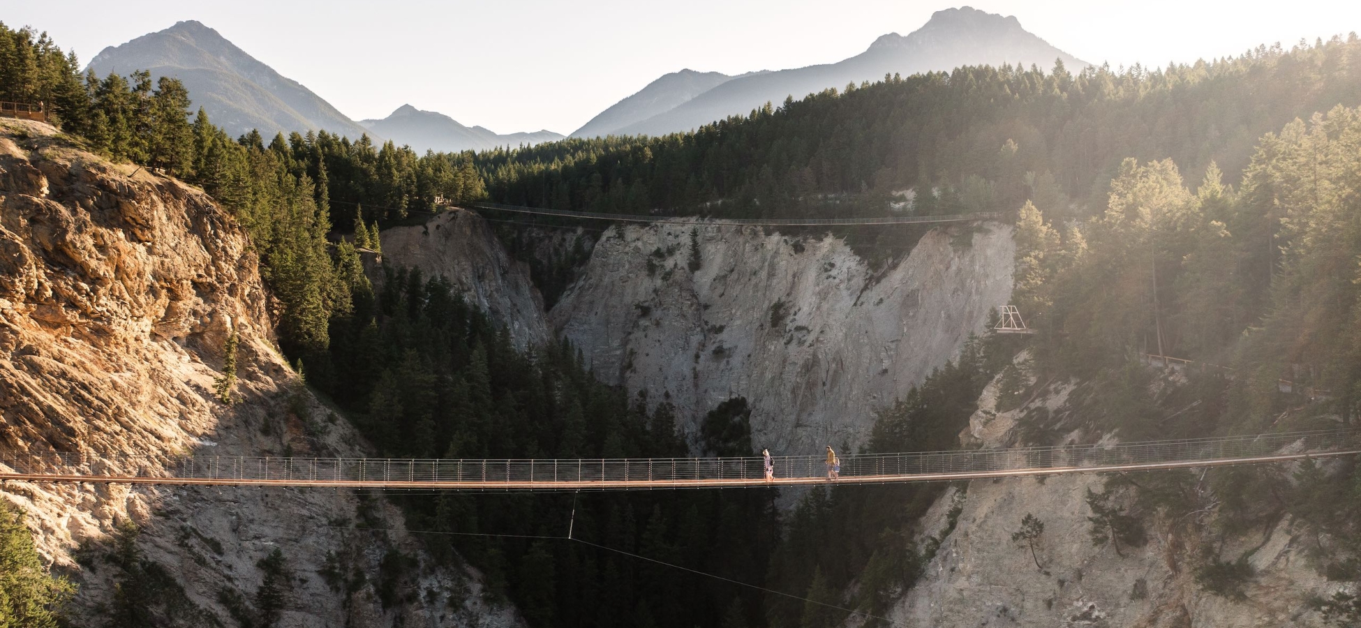 A family of four people walk across the Golden Skybridge between two mountains and over canyon below. There is another suspension bridge behind them in the distance.