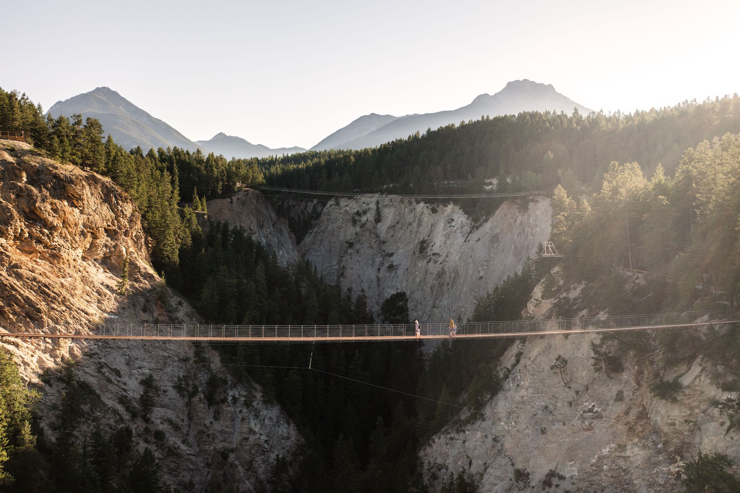 A family of four people walk across the Golden Skybridge between two mountains and over canyon below. There is another suspension bridge behind them in the distance.