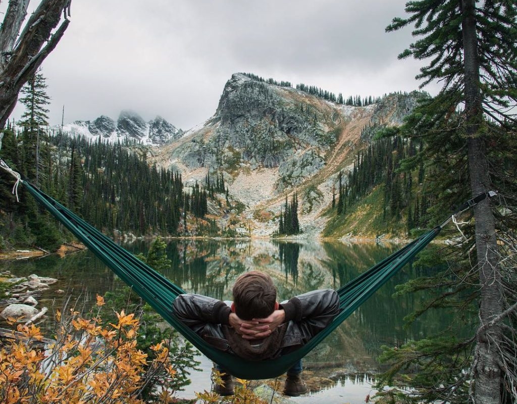 A man relaxes in a hammock overlooking a small pond and a rocky mountain.
