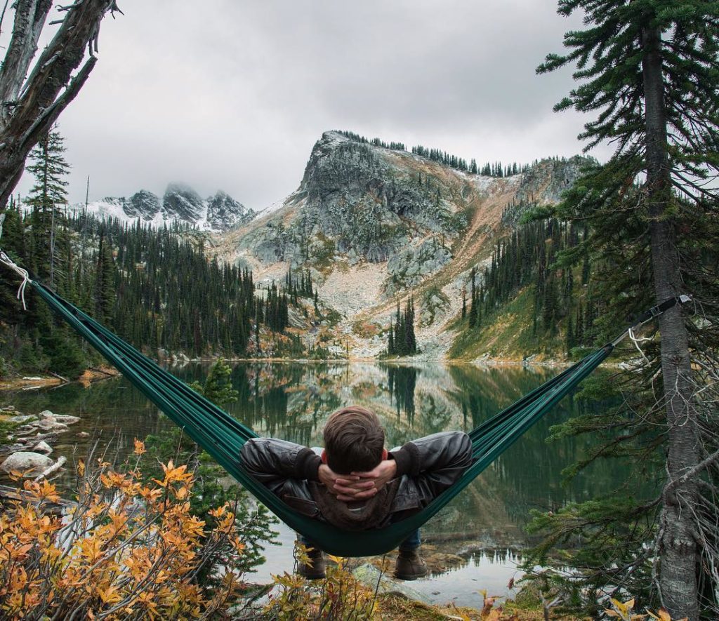 A man relaxes in a hammock overlooking a small pond and a rocky mountain.